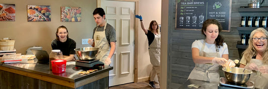 Employees making tea blends and spice blends in a store with woman waving hi in background.