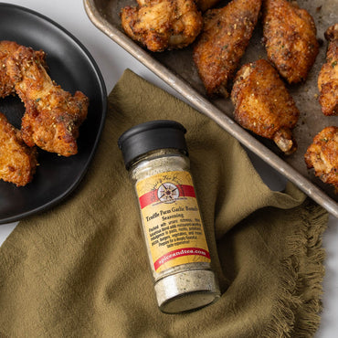 Truffle Parm Garlic Seasoning in flip=top shaker jar next to plate and pan of baked chicken wings and drumstocks